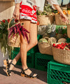 Person wearing black clog sandals with brass stud details holding a woven basket with a colorful background of fruits and vegetables