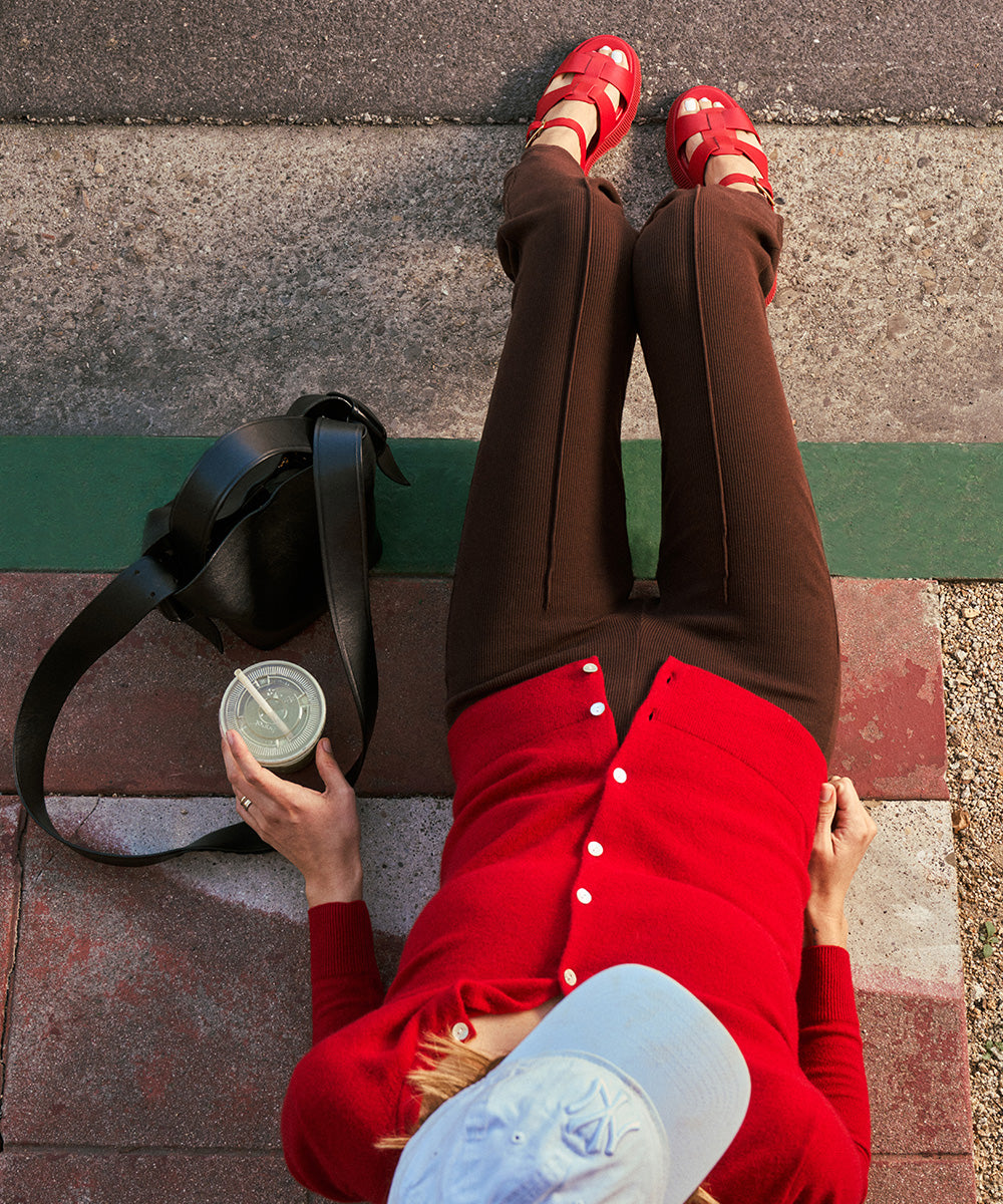 Person wearing red leather fisherman-inspired sandals sitting on a bench with a black bag and a drink.