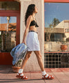Woman wears Henny sandals in Sienna, paired with a black top and white shorts walking outdoors with a blue jacket over her arm.