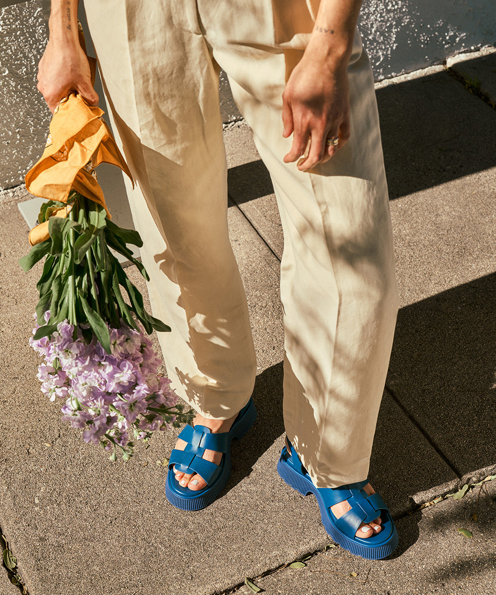 Person wearing blue leather suede platform sandals and beige pants holding a bouquet of flowers on a sidewalk.