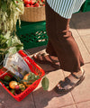 Person wearing brown raffia fisherman sandals  standing on a sidewalk with a red shopping basket containing groceries, next to green crates and plants.