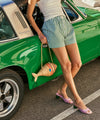 Woman in low heel purple suede sandals with green wooden heel, striped shorts standing next to a green vintage car, holding a straw bag.
