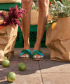 Person wearing cork bed sandals with green and blue suede upper straps standing on a sidewalk next to shopping bags and fruits.