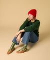 Woman wearing a green sweater, red beanie, blue jeans, and green socks sitting on a beige background