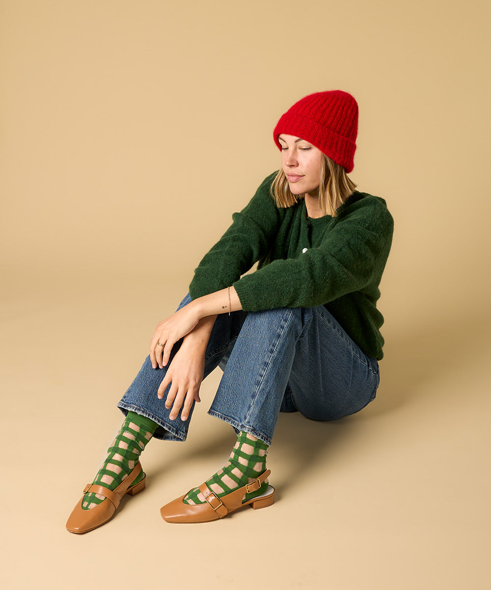 Woman wearing a green sweater, red beanie, blue jeans, and green socks sitting on a beige background